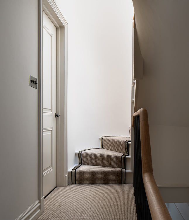 A stylish staircase with a patterned runner, leading up to a white wall, next to a closed door and a wooden banister.
