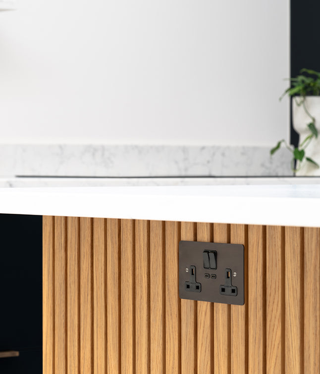 A close-up of a modern kitchen with a wood-paneled base and a dark power outlet against a white countertop with plants in the background.