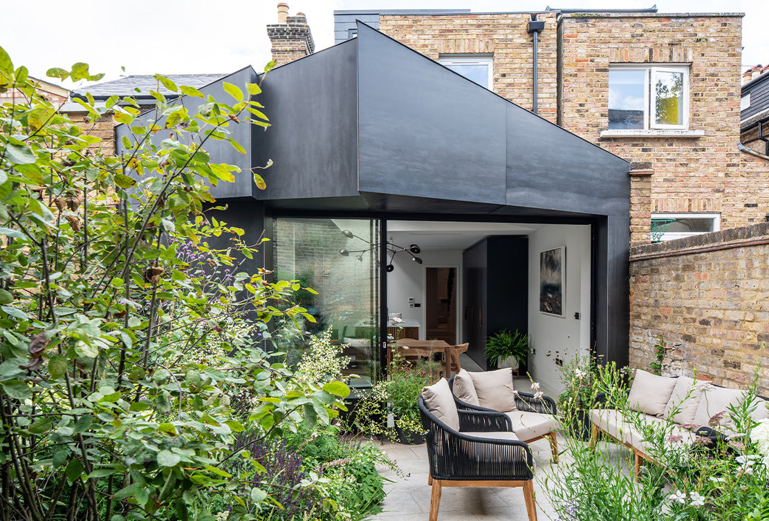 Modern black extension of a home surrounded by lush greenery, featuring a cozy seating area with soft cushions and a dining table inside.