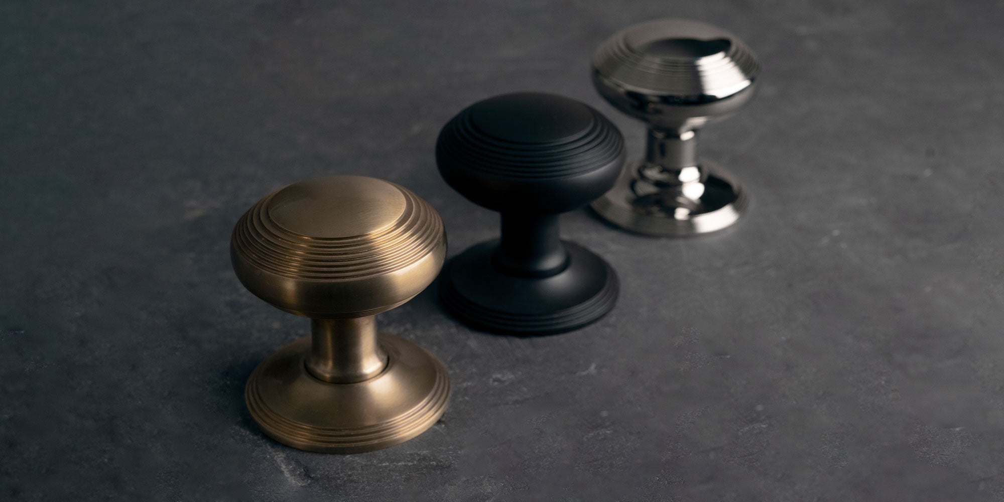 Three decorative cabinet knobs in different finishes: antique brass, matte bronze, and polished nickel, arranged on a dark surface.