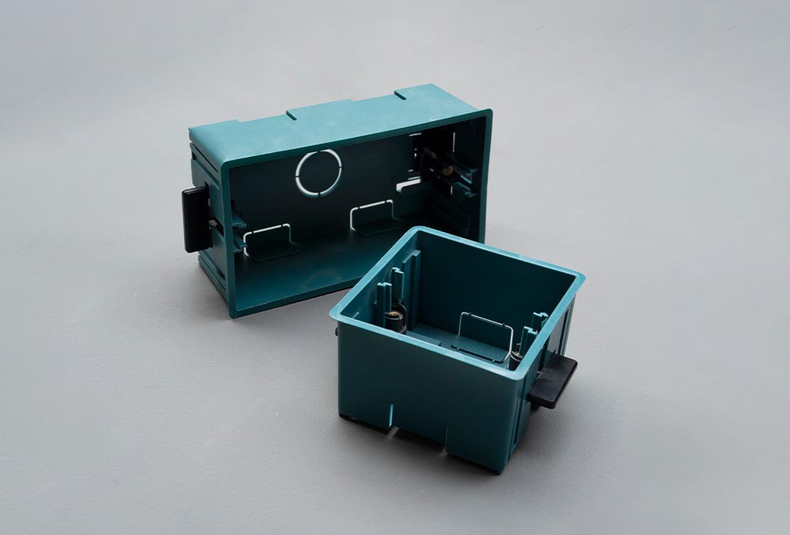 Two teal plastic electrical boxes, one larger and open with a circular hole, next to a smaller, closed box. Both feature internal clips.