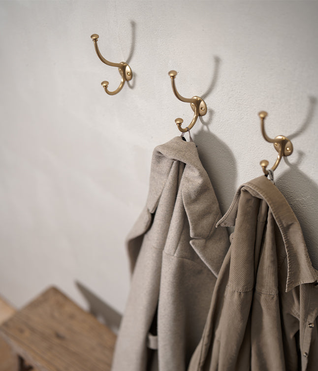 Two beige coats hanging on elegant brass hooks mounted on a textured white wall, with a rustic wooden bench in the foreground.