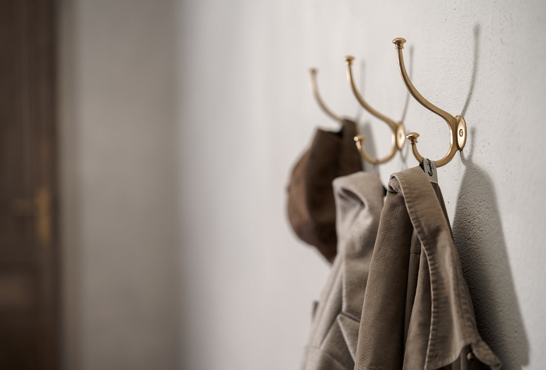 Three golden hooks on a beige wall hold a brown hat and two beige jackets, creating a cozy and stylish entryway look.