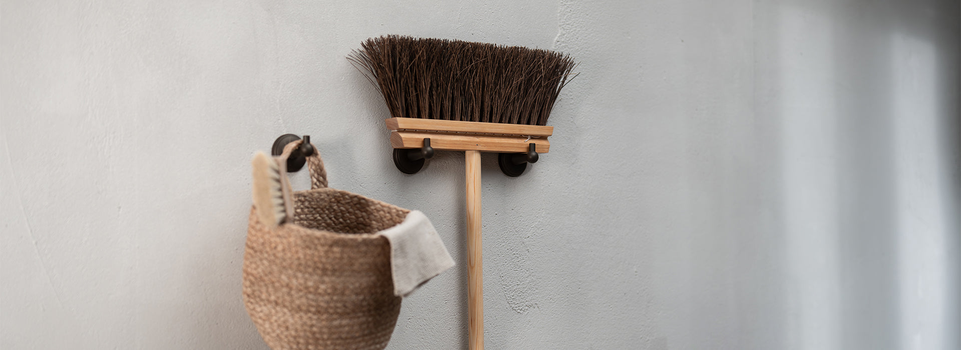 A wooden broom with a natural bristle head hangs on a wall next to a woven basket, showcasing a minimalist cleaning setup.