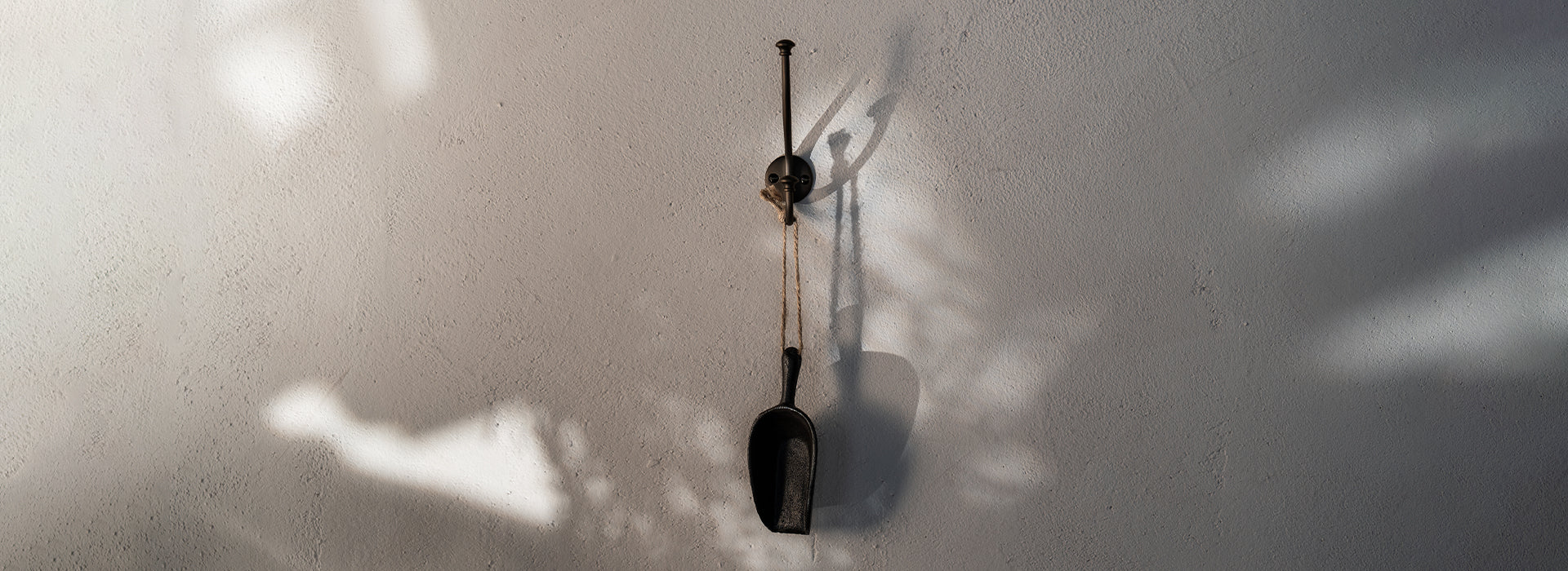 An iron scoop hangs from a black hook on a textured white wall, casting subtle shadows from nearby light.