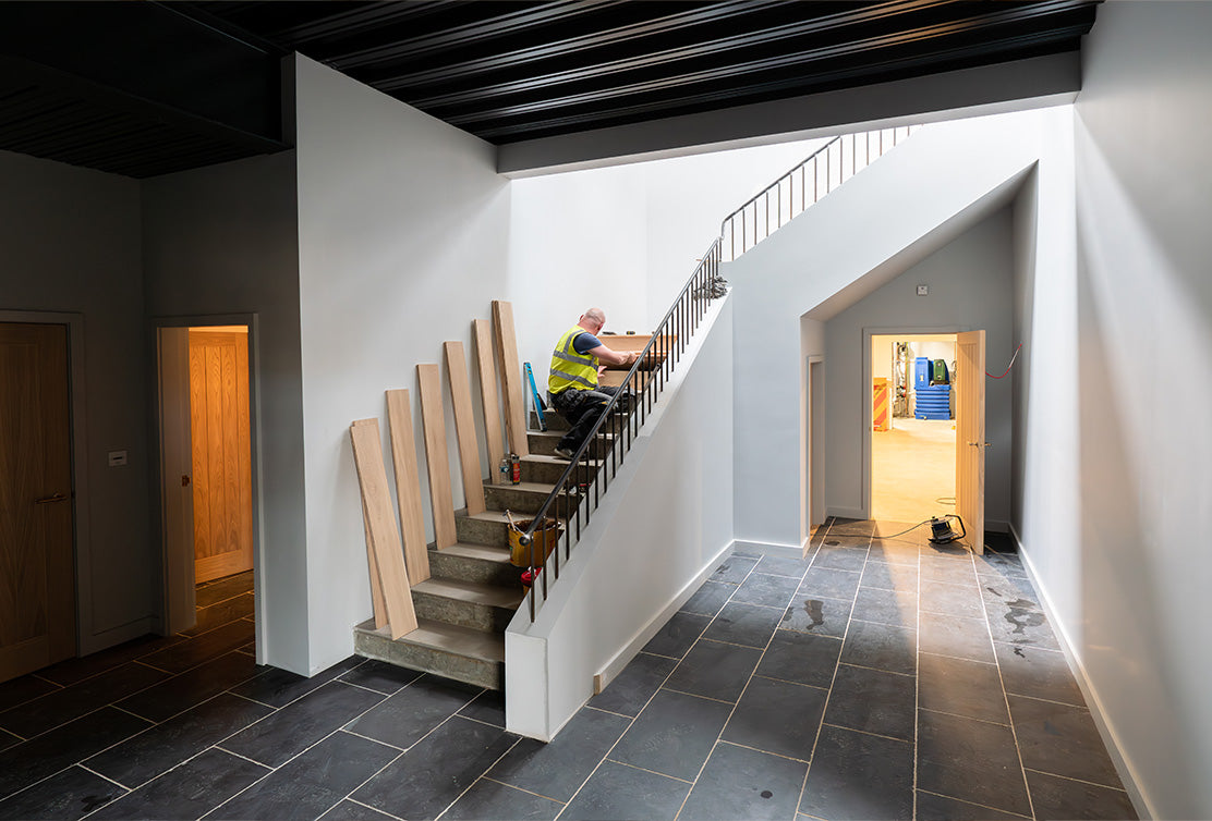A worker in a yellow vest installs a railing on a staircase in a modern, bright hallway with gray tile flooring.