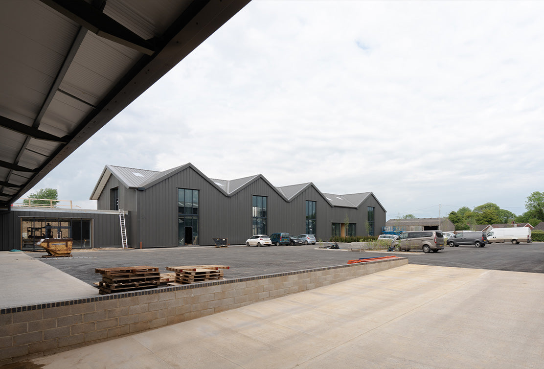 View of modern industrial buildings with sloped roofs, surrounded by parked vehicles and construction materials under a cloudy sky.