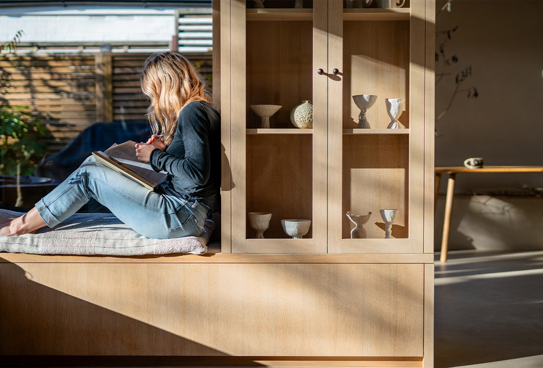 Sunlit room with a person sitting in a window nook. Storage cupboard with glass panes on the doors