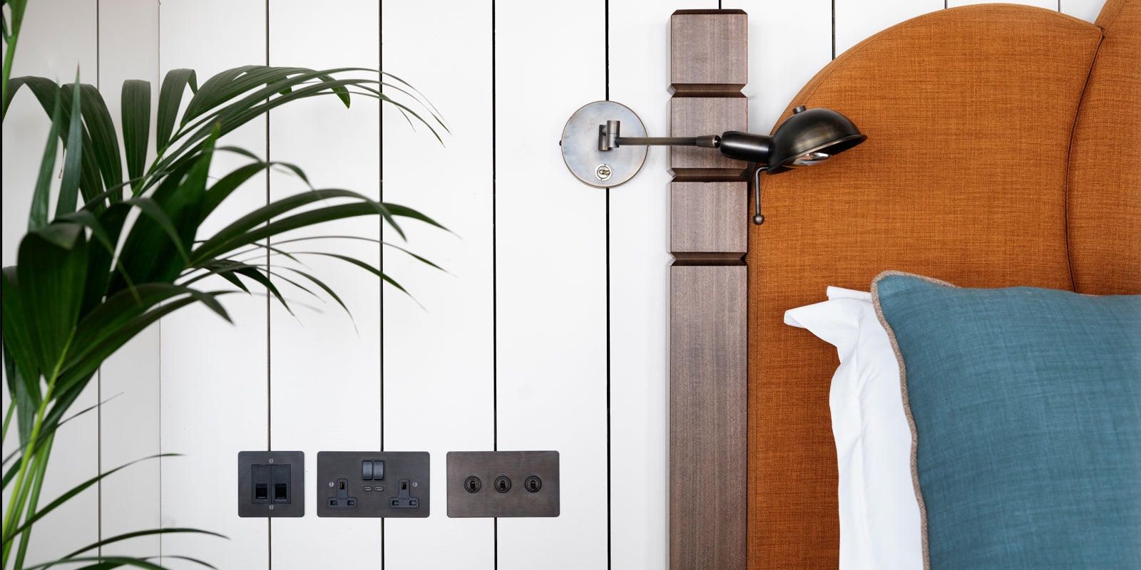 A cozy bedroom corner featuring a stylish orange upholstered headboard, a wall-mounted lamp, and power outlets, framed by lush greenery.