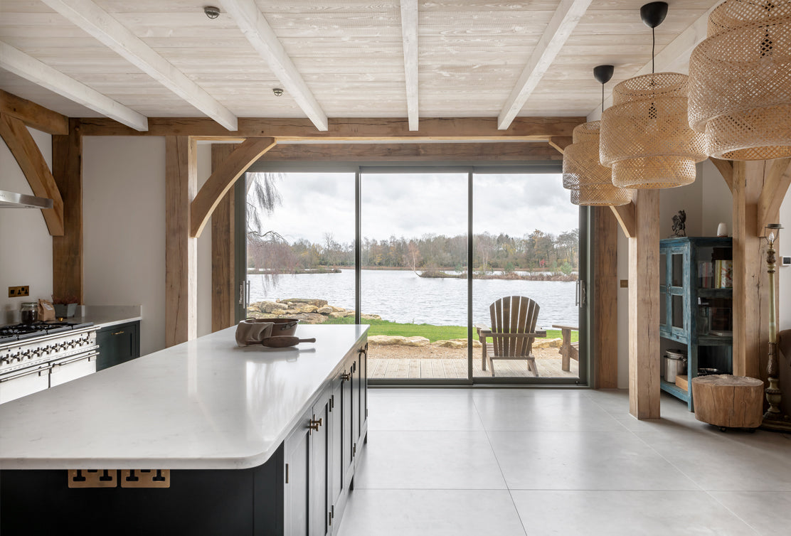 Bright kitchen with a marble island, wooden beams, and lake view through large sliding glass doors. Cozy chairs sit outside.
