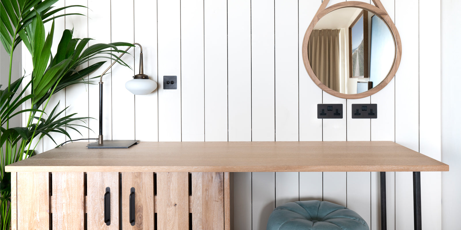 A modern wooden desk with a round mirror, lamp, and potted plant against a white paneled wall, showcasing contemporary decor.