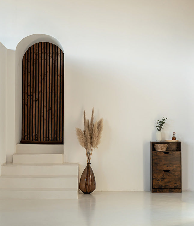 Minimalist interior featuring a wooden arched door, white staircase, pampas grass in a vase, and a rustic drawer cabinet.