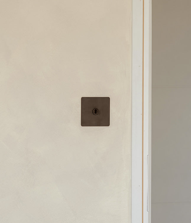 A minimalist black square wall switch set against a textured beige wall, with an adjacent light-colored door frame.