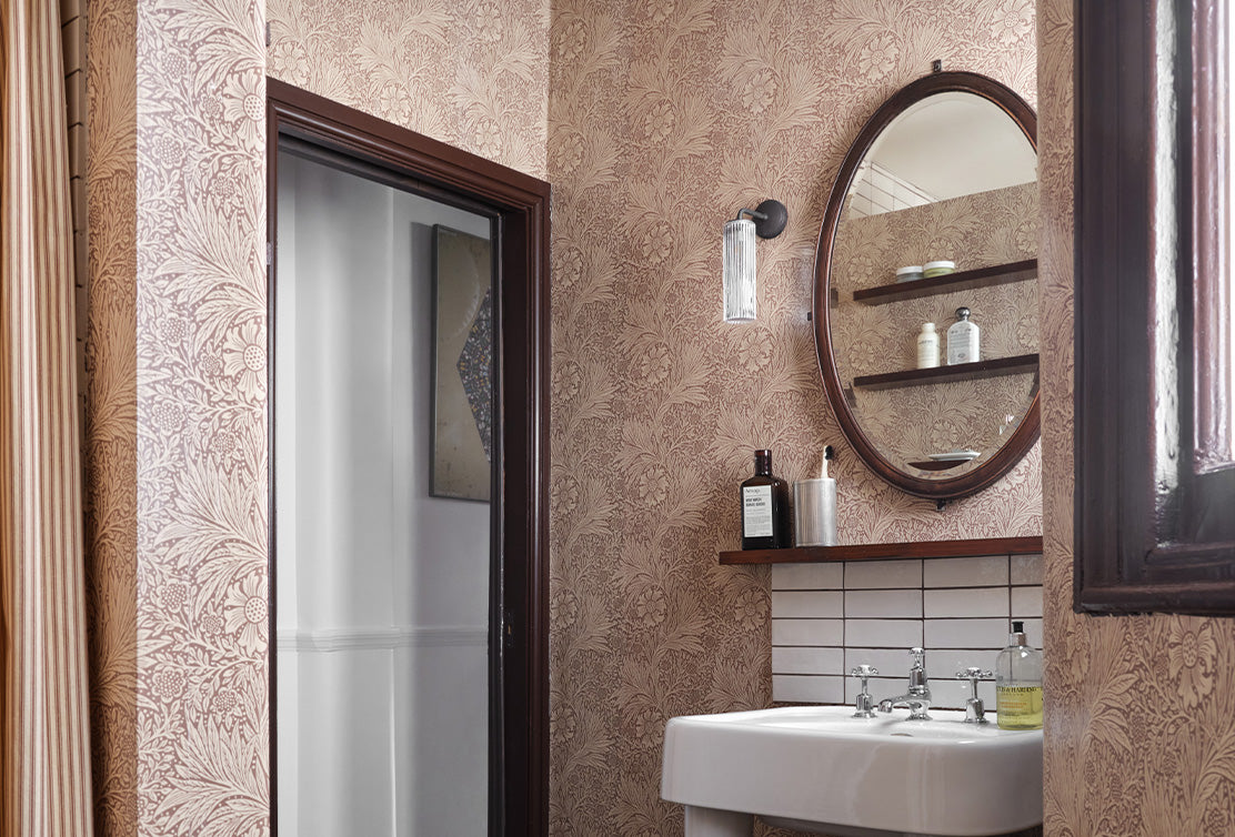 A stylish bathroom featuring floral wallpaper, a round mirror, a glass light fixture, and a minimalist sink with shelving.