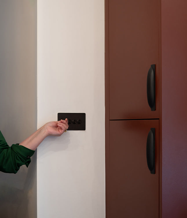 A person's hand adjusts a sleek black light switch next to a modern brown cabinet in a minimalist kitchen setting.