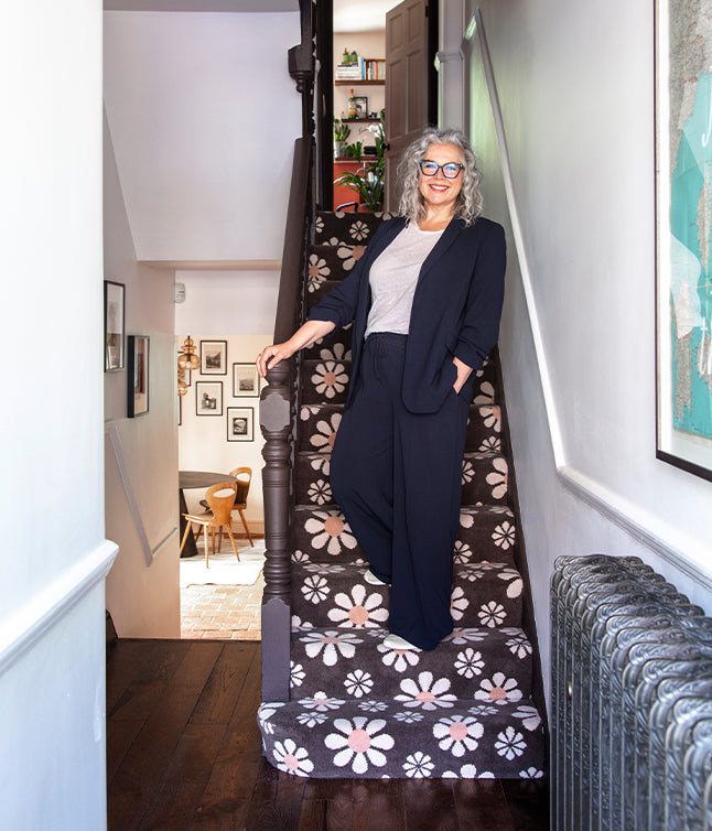 A person in a navy suit and grey top stands on a flower-patterned staircase, leading to a stylish, light-filled room.