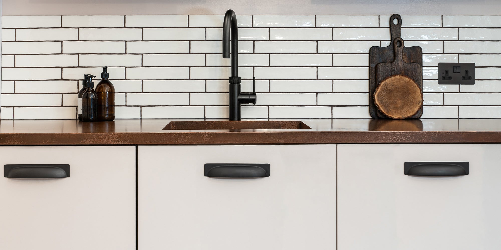 Modern kitchen countertop with a black faucet, glass bottles, wooden cutting board, and white subway tile backsplash.