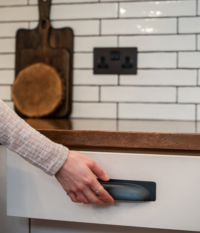 A hand reaches for a sleek black drawer handle in a modern kitchen with white tile backsplash and wooden cutting board in the background.