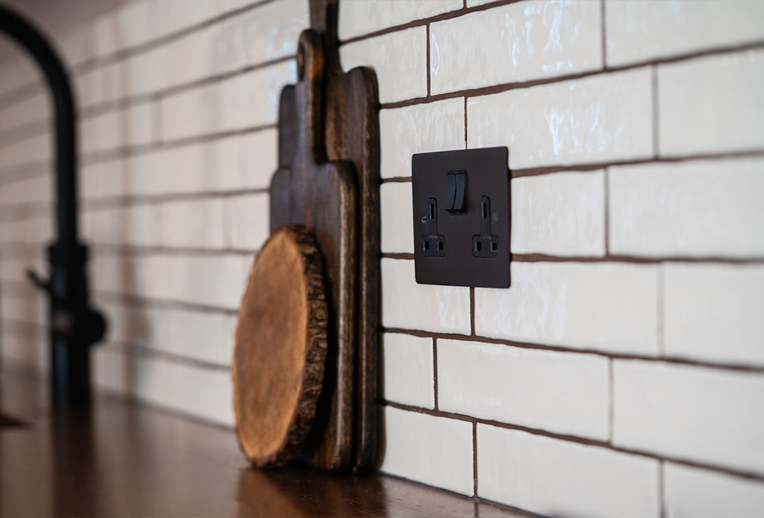 A dark electrical outlet on a white tiled kitchen wall, beside a wooden cutting board with a natural edge.