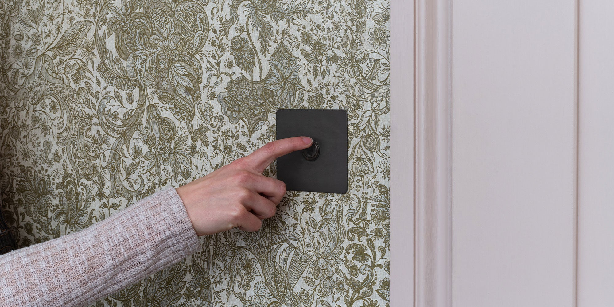 A hand presses a black switch against a decorative floral wallpaper, adding a modern touch to the vintage-inspired design.
