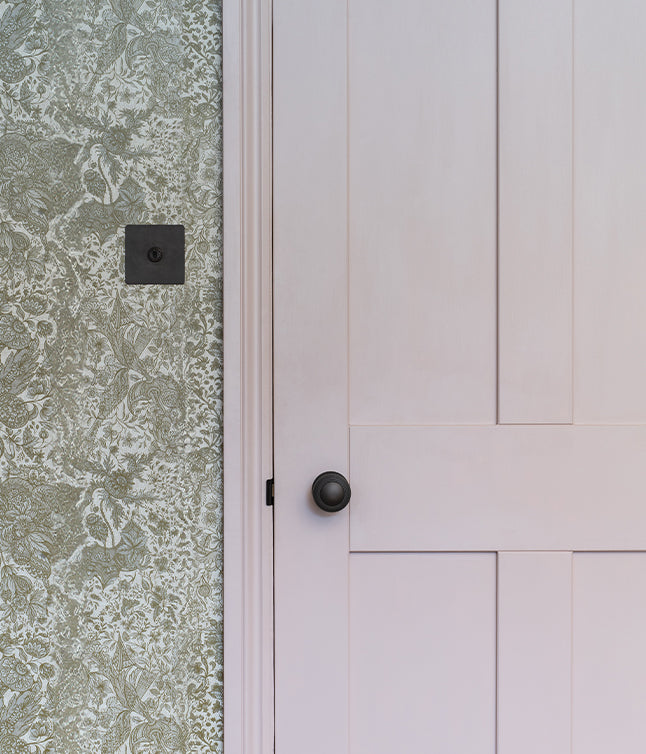 A close-up of a pale pink door with a black knob, adjacent to textured green floral wallpaper.