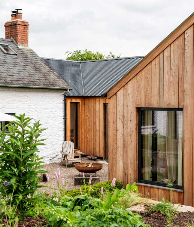 A modern wooden extension connects to a traditional house, featuring a fire pit and a comfortable seating area surrounded by lush greenery.