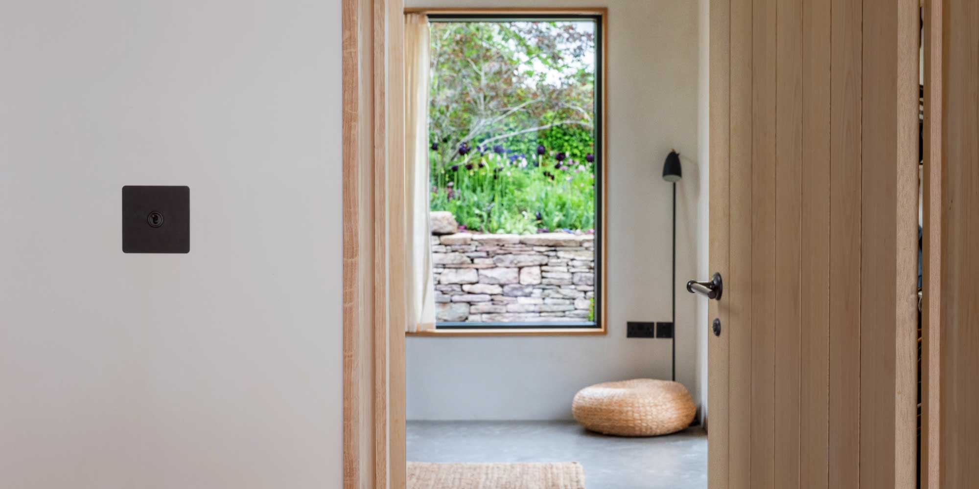 A glimpse through an open door, revealing a lush garden view framed by a stone wall and a cozy, woven pouf in the foreground.