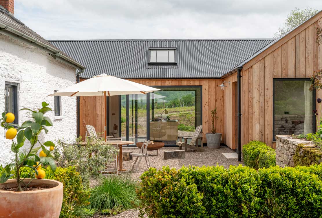 A modern cottage with a wooden exterior, featuring an outdoor seating area under an umbrella, surrounded by greenery and a scenic landscape.