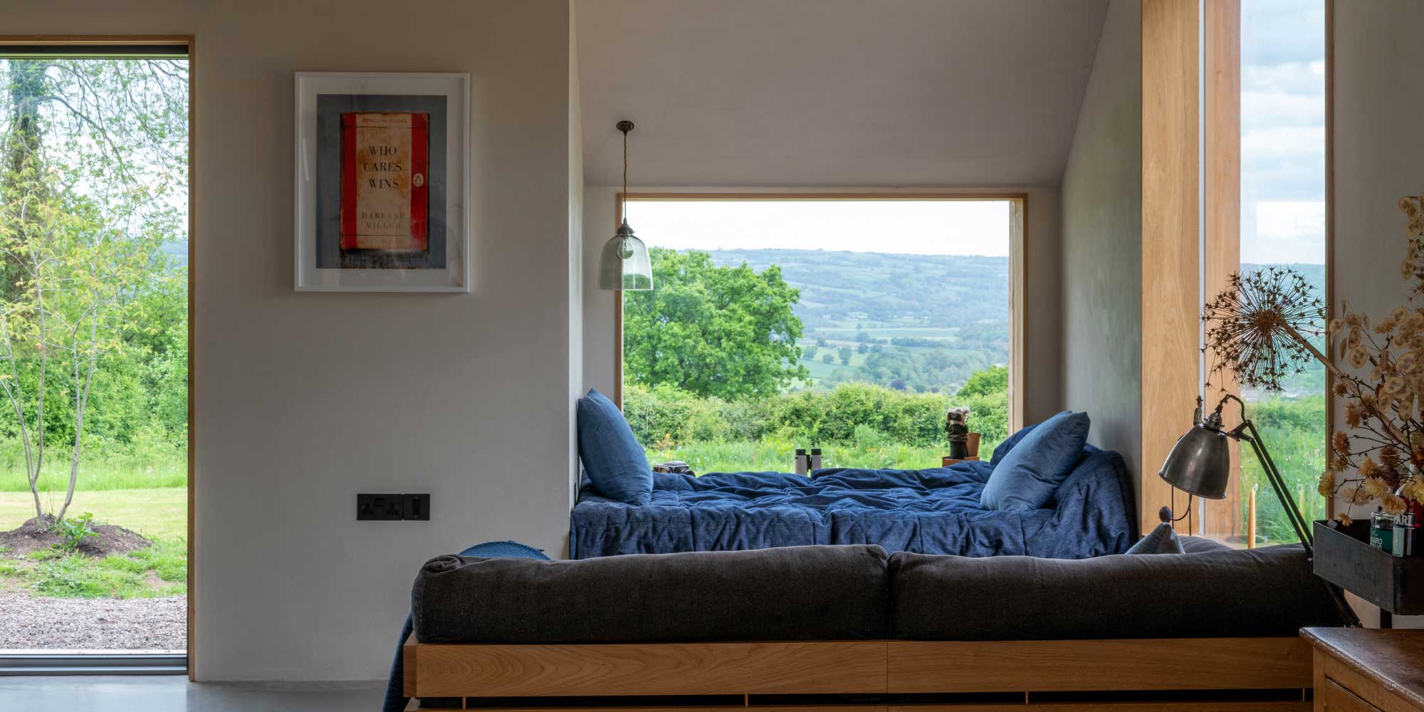 Cozy bedroom with two blue beds, a framed book cover on the wall, and a large window showcasing a scenic countryside view.