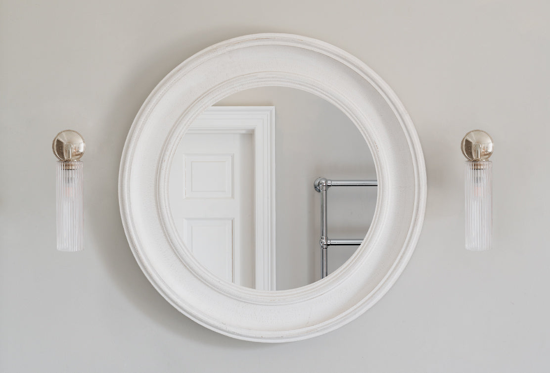 A round white-framed mirror surrounded by stylish polished nickel and glass wall sconces, set against a neutral grey wall.