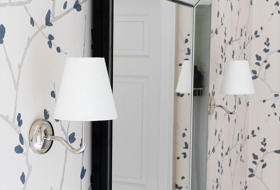 A modern wall sconce with a white shade is mounted on a light floral patterned wall, reflecting in a nearby mirror.