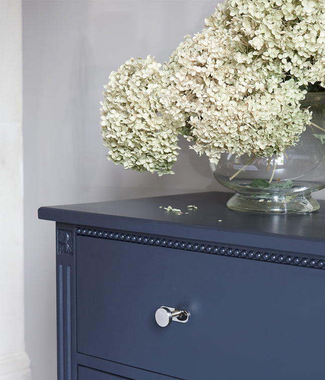 A navy dresser topped with a large glass vase filled with pale dried hydrangeas, against a soft gray wall.
