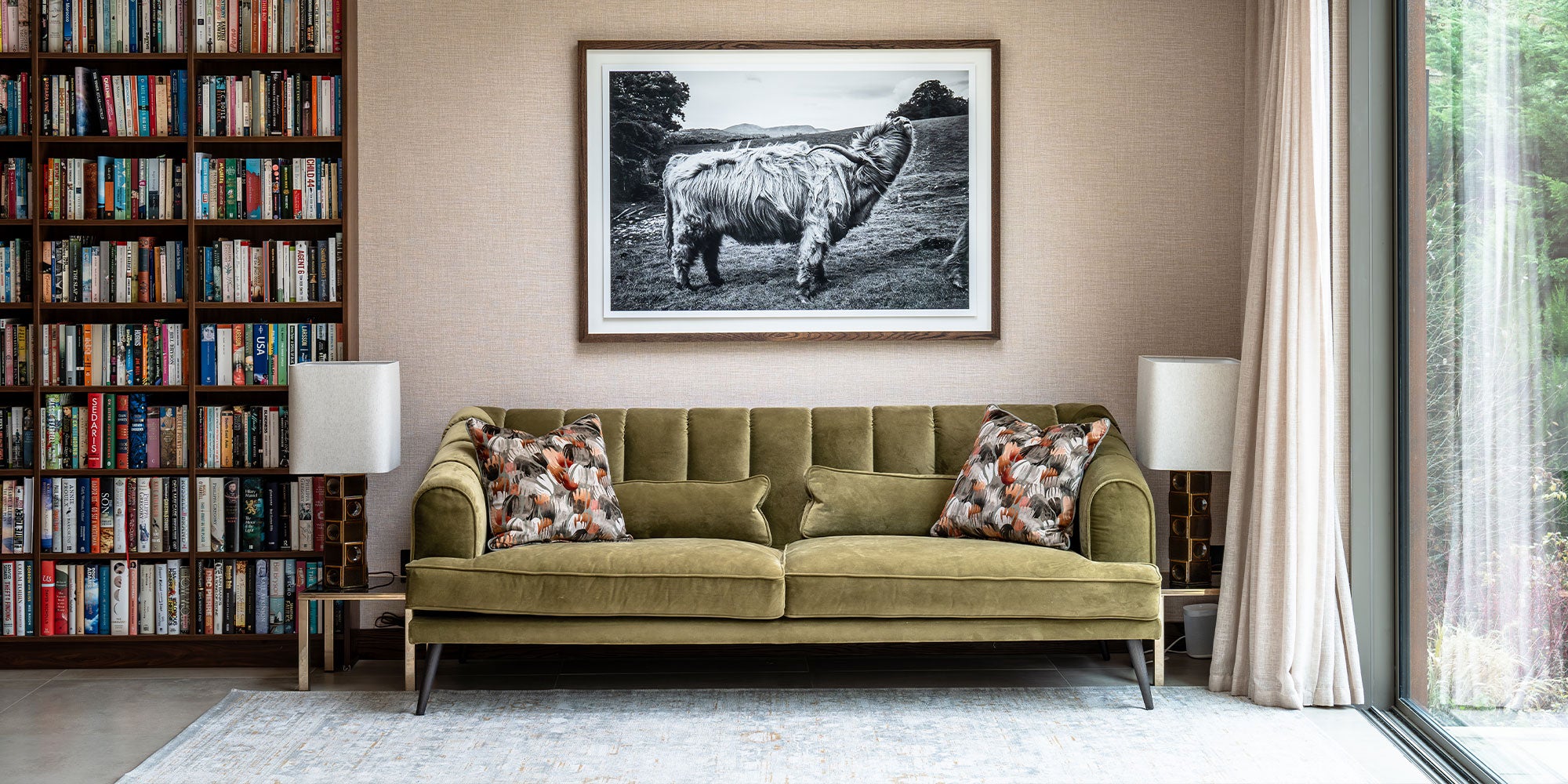 A stylish living room featuring a green velvet sofa with patterned pillows, a lamp, a wall of books, and a black-and-white cow photograph.