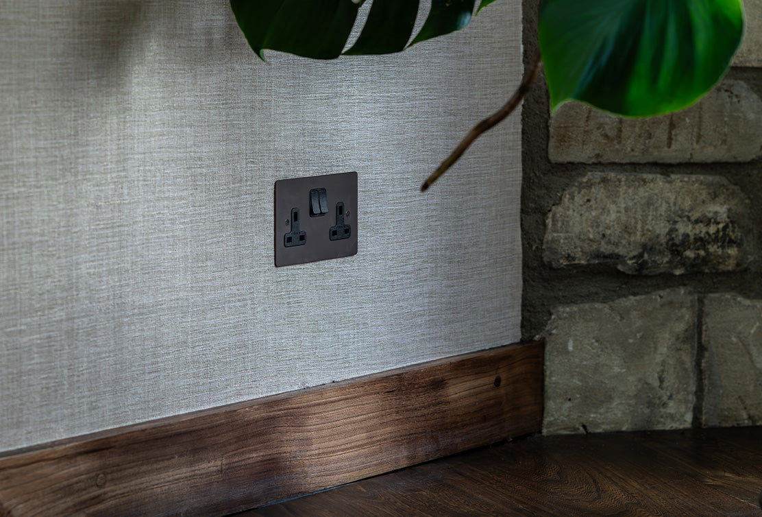 A dark electrical outlet with dual sockets and a switch, set against textured wallpaper and wooden trim, beside a stone wall.