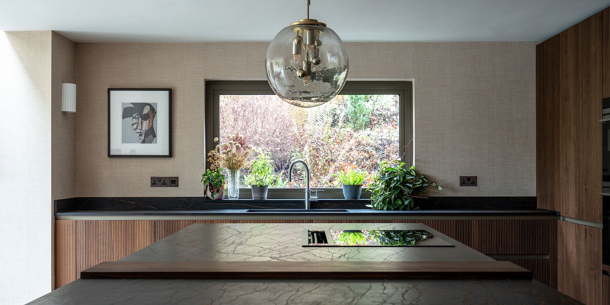 A modern kitchen with wood accents, a large window view of greenery, and a stylish pendant light, featuring a bowl and potted plants.