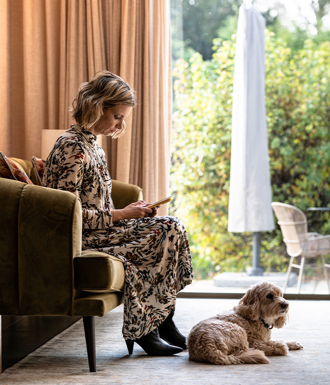A woman in a floral dress sits on a green sofa with a phone, while a small dog relaxes beside her on a plush rug.