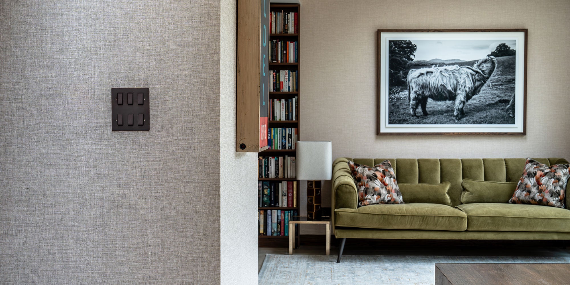 Modern living room featuring a green velvet sofa with patterned pillows, a bookshelf, and a large black and white cow photograph on the wall.