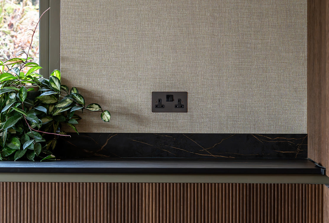 A textured beige wall with a black marble countertop, featuring a power outlet and lush green leaves from a potted plant.