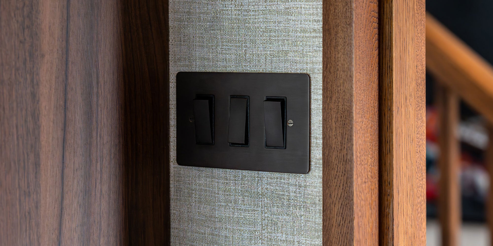 A close-up of a modern, black three-switch light panel against a textured gray wall and wooden trim.