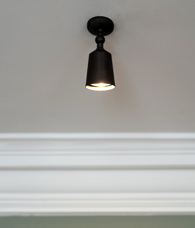 A close-up of a lit bronze 'Baylis Single' spotlight which is angled directly downwards towards the floor.