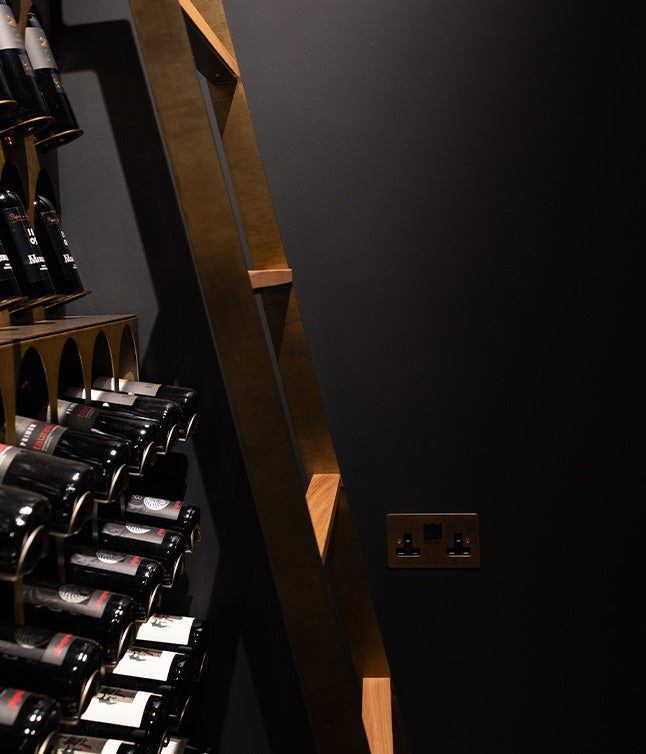 A wooden ladder against a dark wall, beside a wine rack filled with bottles and a modern electrical outlet.