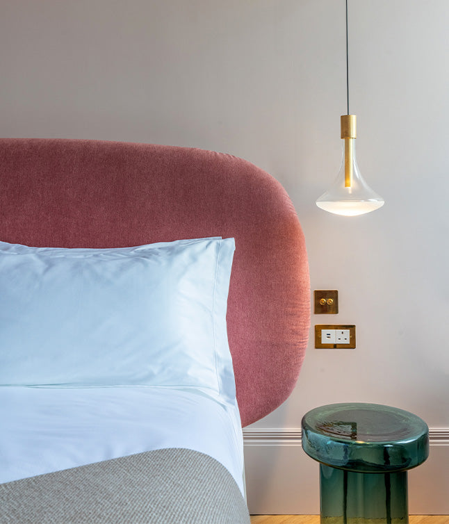 Modern bedroom featuring a pink upholstered headboard, white bedding, a green side table, and a stylish pendant light.