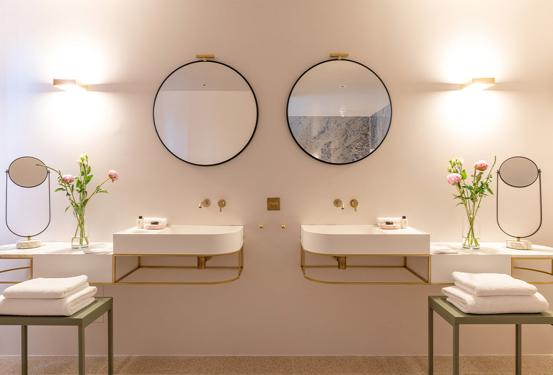 Modern bathroom with dual sinks, circular mirrors, warm lighting, and fresh flowers on elegant countertops, featuring white towels.