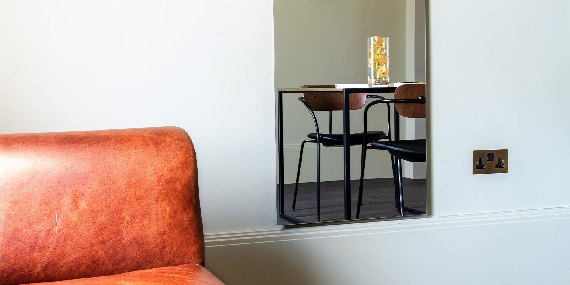 Close-up of a leather sofa with a mirror reflecting a small dining table and chairs, accented by a decorative vase with yellow flowers.