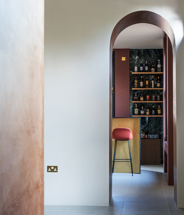 Modern interior with an arched doorway, sleek bar area, and a red stool in front of a dark green marble shelf stocked with liquor.