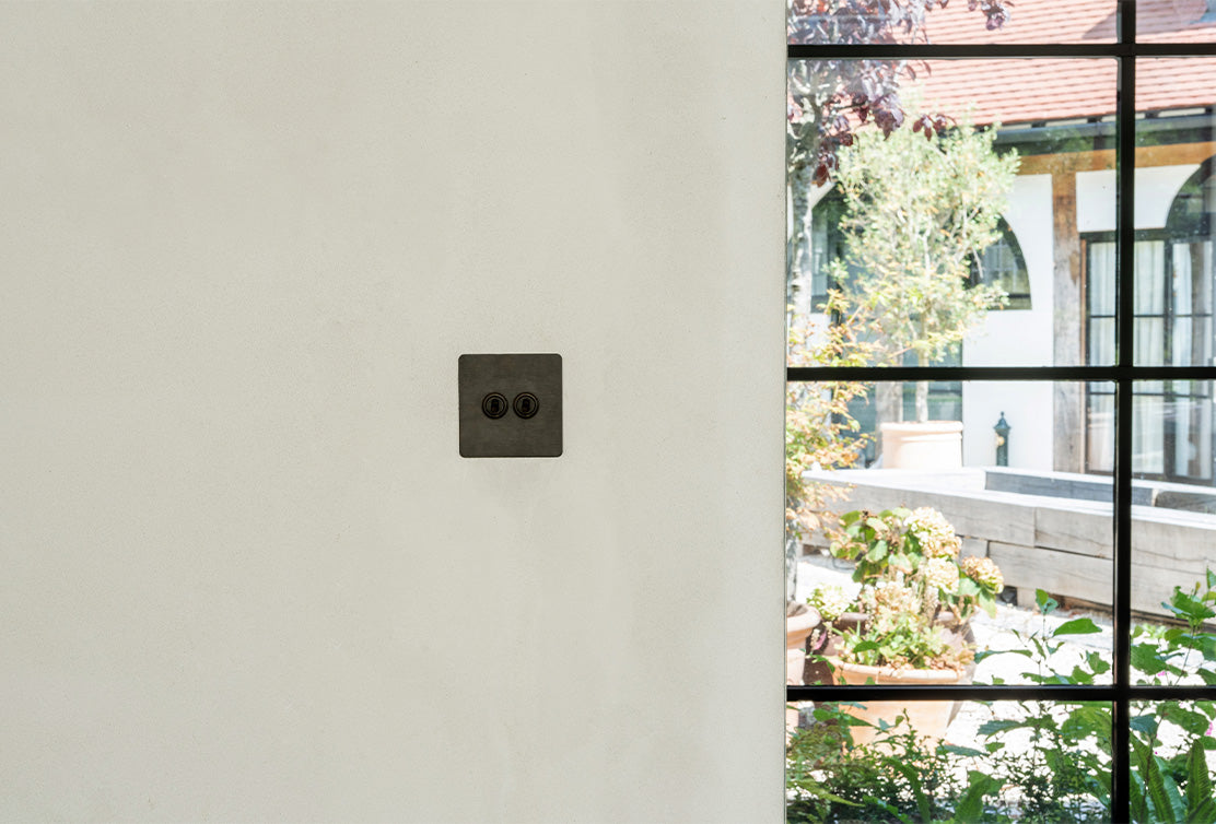 A close-up of a modern black electrical outlet on a smooth white wall, with a garden visible through a large window beside it.