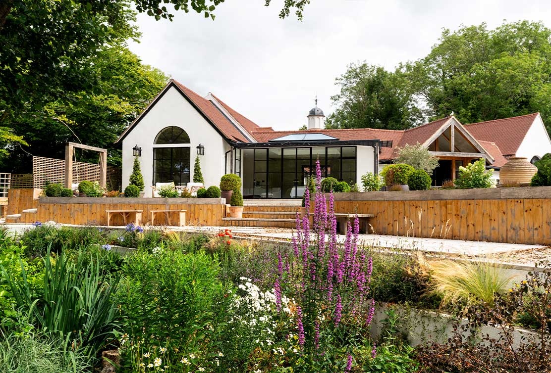 A modern white house with large glass windows, surrounded by landscaped gardens and wooden decking, under a cloudy sky.