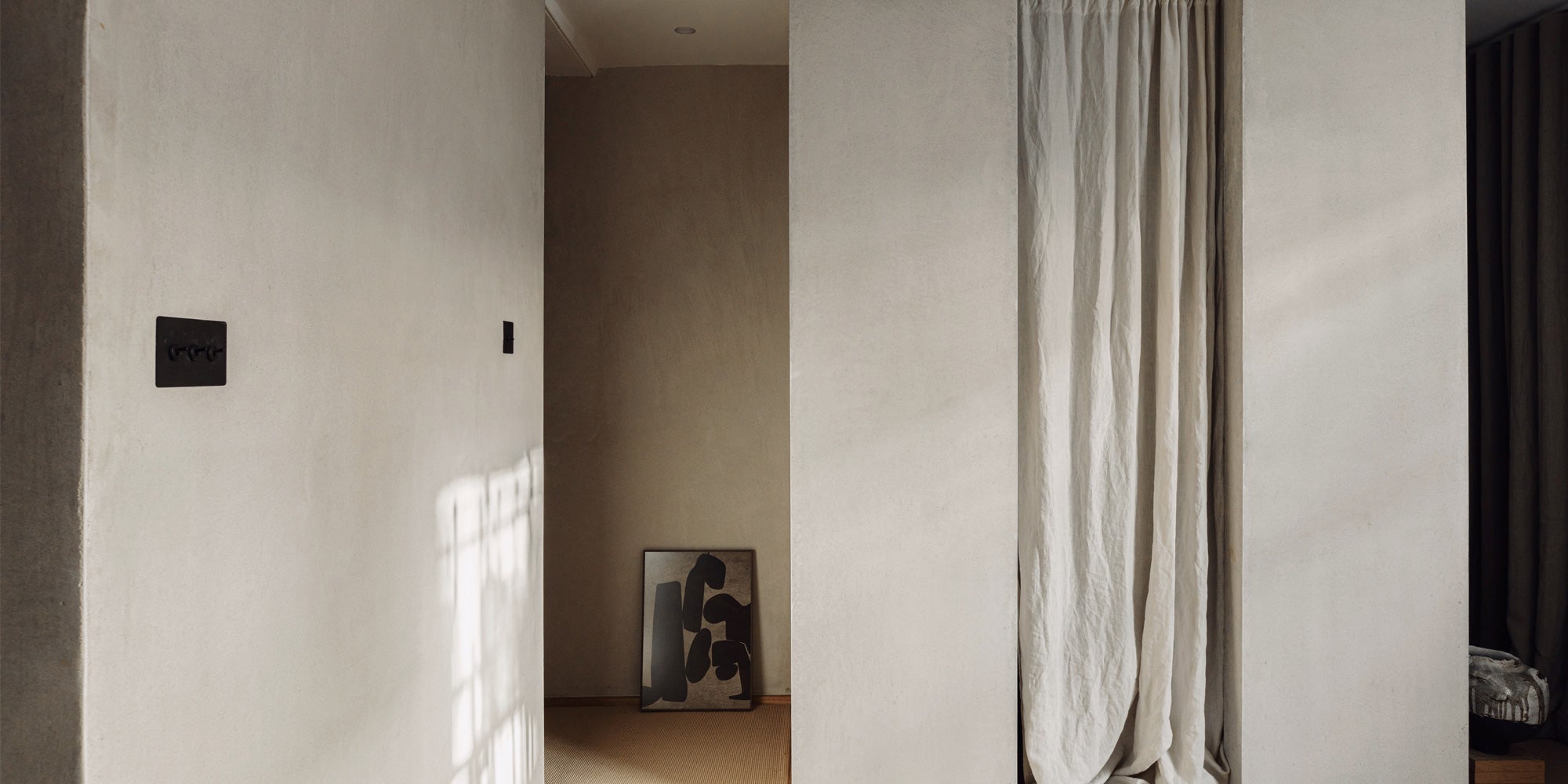 A neutral-toned hallway features a textured wall, a black power switch, and an abstract black artwork against a soft beige background.