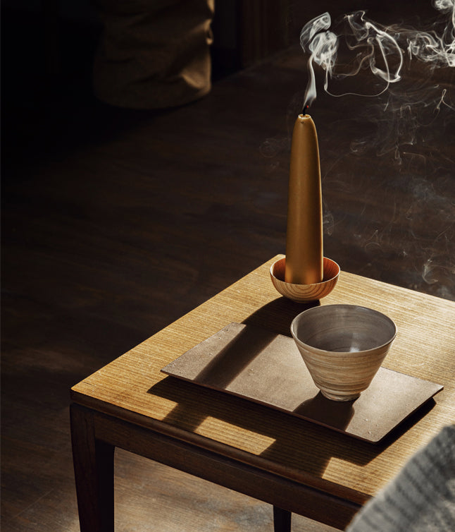 A golden candle burns on a wooden table, with smoke wafting above two ceramic bowls and a leather mat, casting soft shadows.
