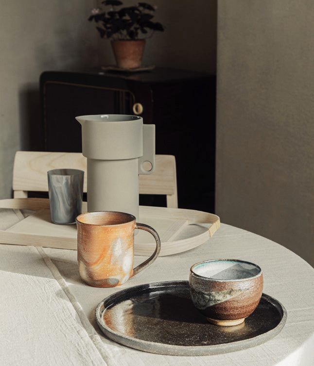 A cozy table setting featuring a gray pitcher, two artisanal cups, and a round tray on a textured linen tablecloth.